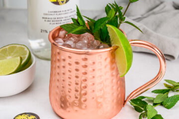 A copper mug filled with crushed ice, garnished with mint leaves and a lime wedge, sits on a counter next to sliced lime and a bottle of coconut rum.
