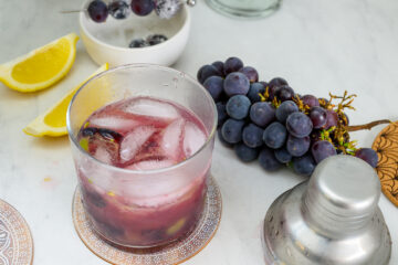 A glass of purple grape cocktail with ice on a table, next to a bunch of grapes, lemon wedges, a cocktail shaker, and a small bowl with grapes.