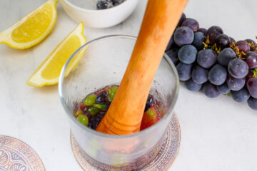 A wooden muddler crushing grapes in a glass, with lemon wedges, a bowl of salt, and a bunch of black grapes on a light surface.