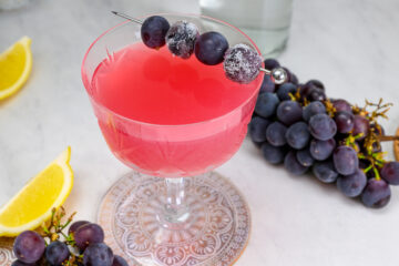 A pink cocktail in a glass garnished with sugared grapes on a skewer, surrounded by clusters of grapes and lemon wedges on a marble surface.