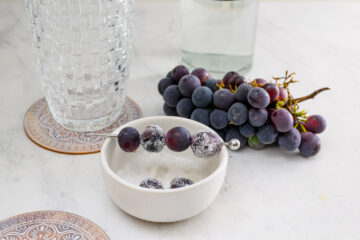 A bunch of grapes sits next to a small white bowl containing sugared grapes and a cocktail pick, with a glass of water and a coaster nearby on a marble surface.