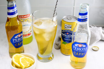 A glass with ice and a yellow drink is surrounded by Peroni beer bottles, a can of Limonata, a bottle of liqueur, and a bowl of lemon slices on a counter.