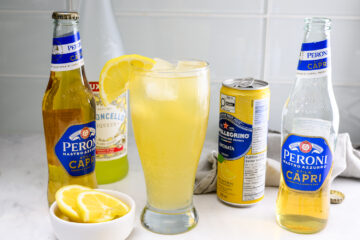 A glass of yellow cocktail with a lemon slice garnish, surrounded by bottles of Peroni beer, lemon liqueur, San Pellegrino Limonata, and a small bowl of lemon slices.
