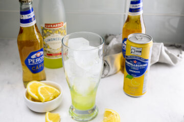 A glass with ice and green liquid sits on a counter beside lemon slices, bottles of Peroni, limoncello, and a can of San Pellegrino Limonata.