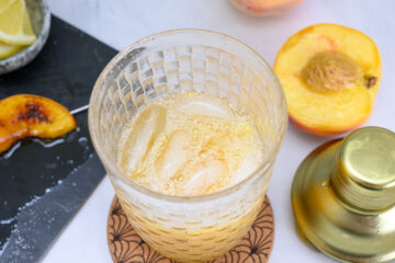 A glass of iced peach drink sits on a coaster next to a halved peach, a gold shaker, and a slate with peach and lime slices.