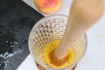 A wooden muddler crushes yellow fruit, likely peach, in a textured glass, with a sliced black cutting board and whole peach nearby.