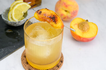 A glass of peach cocktail with ice, garnished with a grilled peach slice on a skewer. Fresh peach halves and lemon slices are in the background on a marble surface.