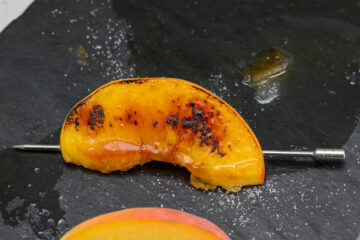 A charred peach slice on a metal skewer rests on a black slate surface, with scattered salt crystals and a small pool of juice nearby.