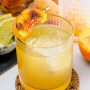 A glass of whiskey cocktail with ice, garnished with a grilled peach slice on a skewer, sits on a coaster with sliced lemon, a peach, and a whiskey bottle in the background.