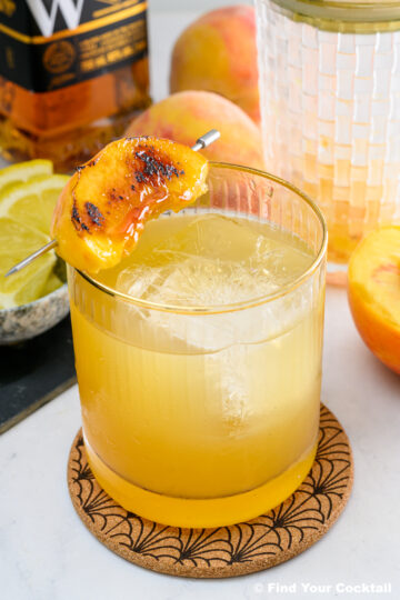 A glass of whiskey cocktail with ice, garnished with a grilled peach slice on a skewer, sits on a coaster with sliced lemon, a peach, and a whiskey bottle in the background.