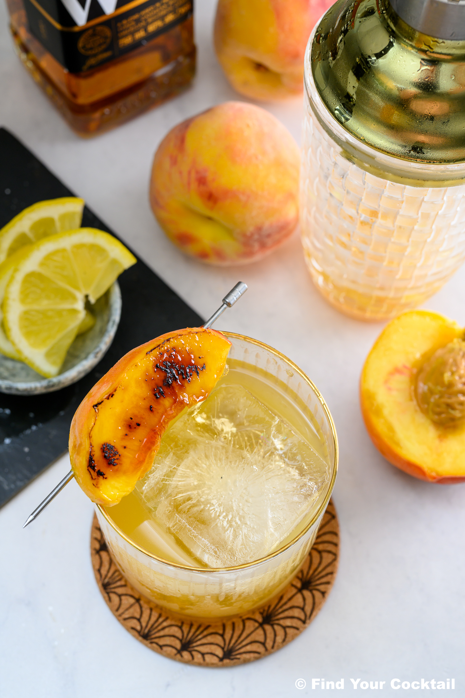 A cocktail with ice and a grilled peach garnish sits on a coaster, surrounded by lemon slices, fresh peaches, a cocktail shaker, and a bottle of whiskey.