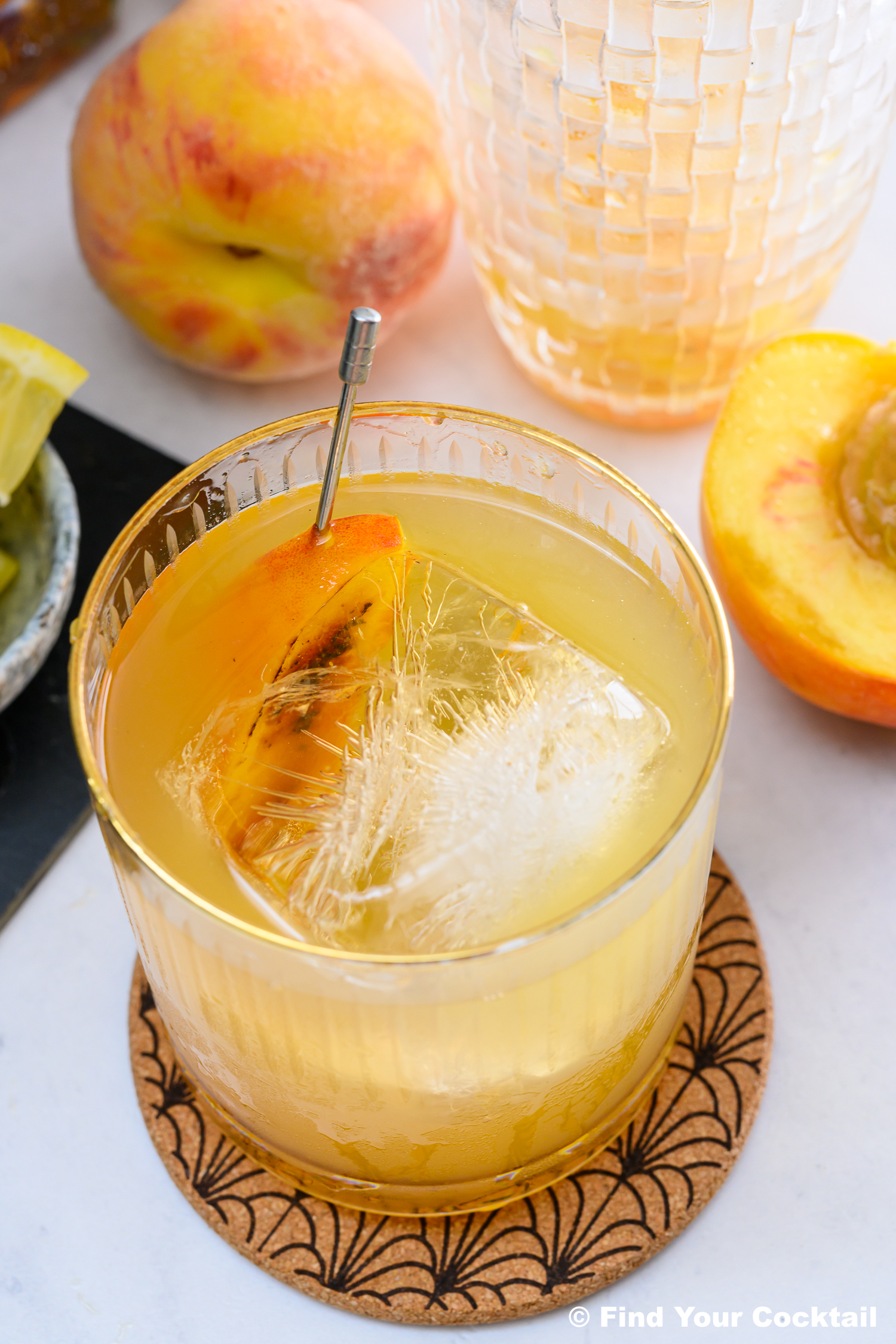 A glass of peach-colored cocktail with a large ice cube and peach slice garnish, placed on a coaster with fresh peaches and a pitcher in the background.