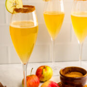 Three champagne flutes filled with a golden drink, garnished with apple slices and cinnamon-sugar rims, are placed among fresh apples and a bowl of brown sugar.