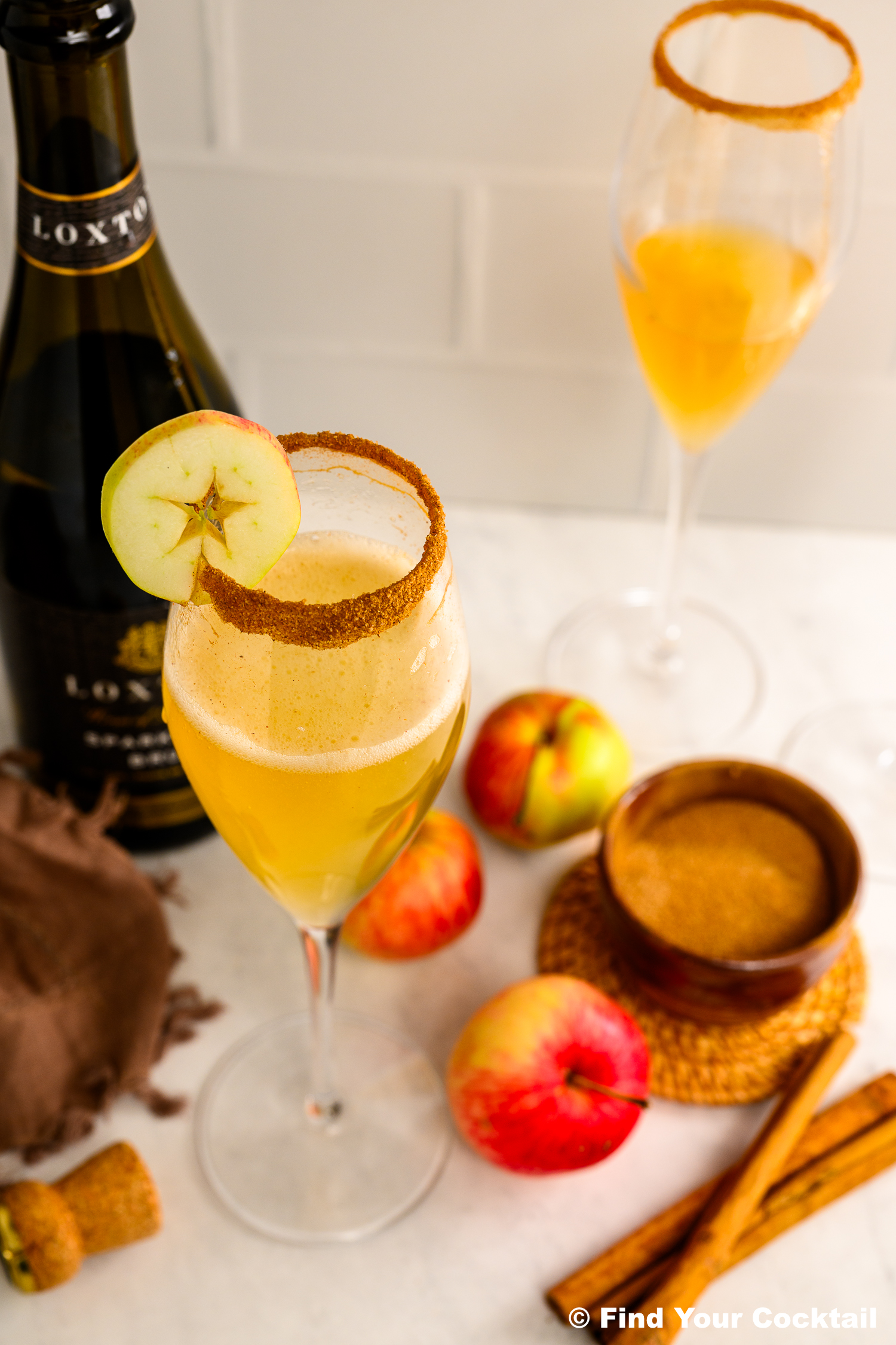Two champagne flutes with golden cocktails, garnished with apple slices and cinnamon-sugar rims. A bottle, apples, cinnamon sticks, and a bowl of brown sugar are on the table.