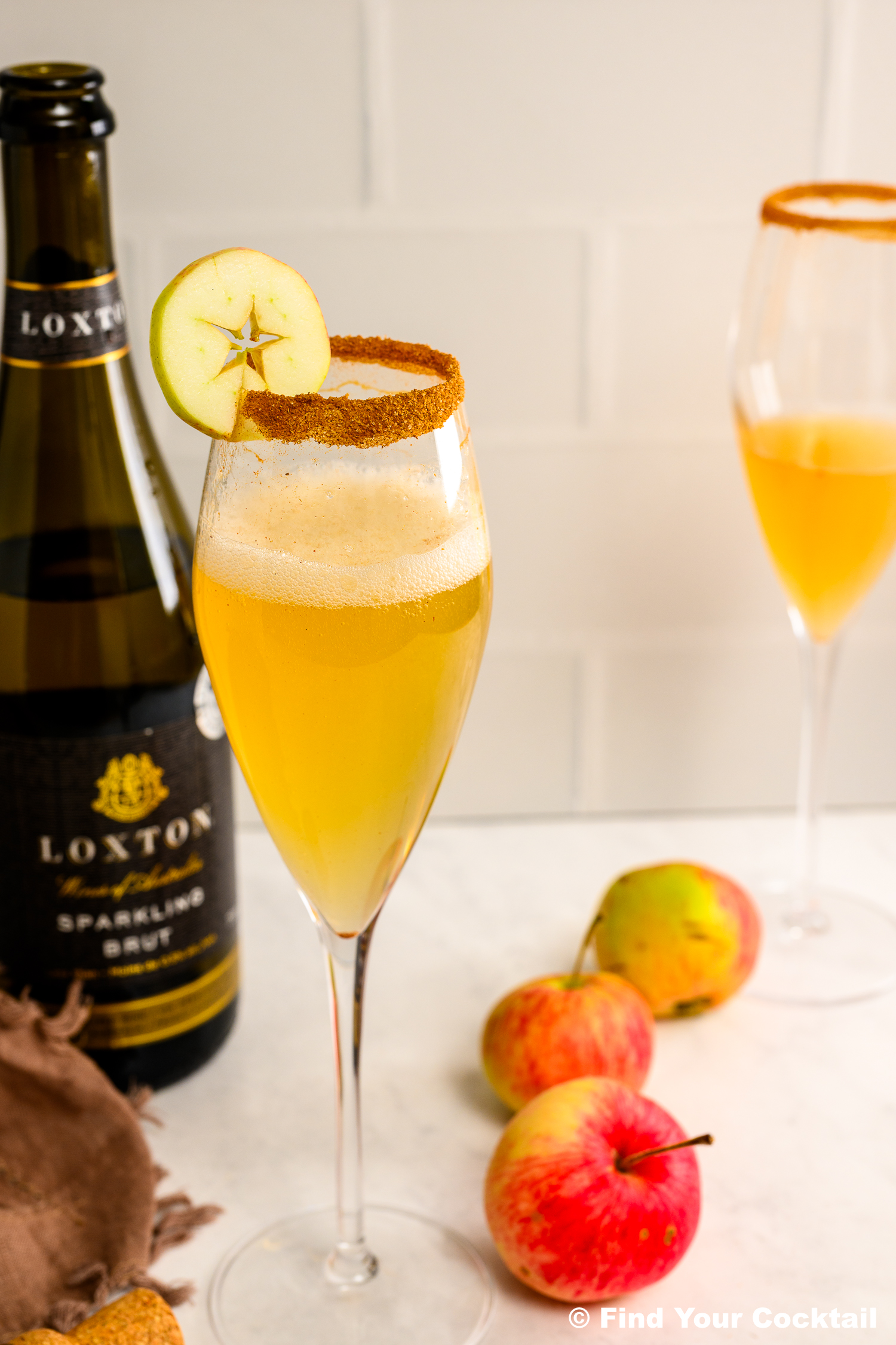 A champagne flute with a spiced rim and apple slice garnish, filled with a yellow cocktail, sits beside apples and a bottle of sparkling brut.