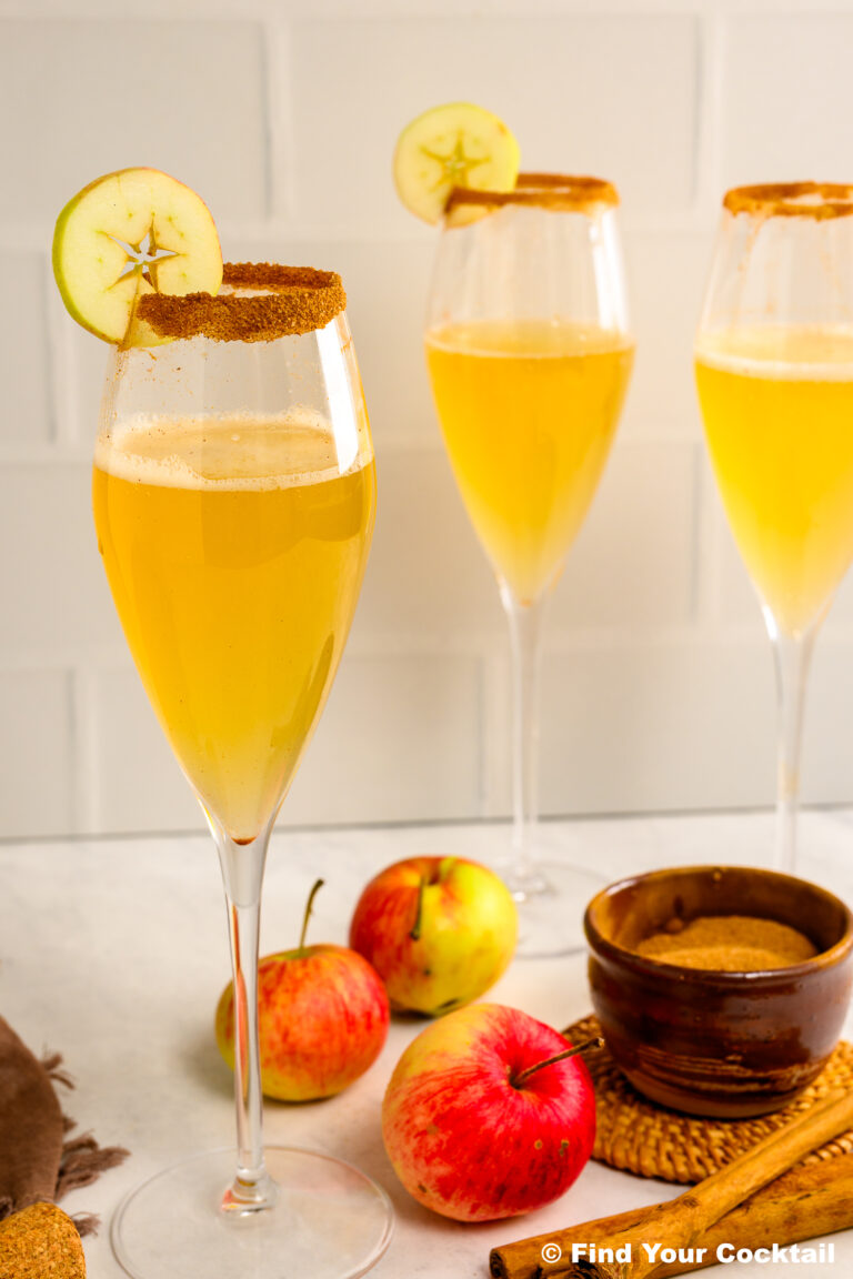 Three champagne flutes filled with a golden drink, garnished with apple slices and cinnamon-sugar rims, are placed among fresh apples and a bowl of brown sugar.