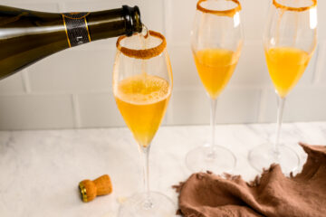 A bottle pours sparkling wine into a champagne flute with an orange drink and a sugared rim; two similar filled glasses are beside it on a marble surface.