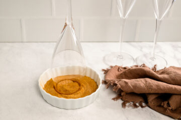 A champagne glass is turned upside down in a dish of cinnamon sugar, with two empty glasses and a brown cloth nearby on a marble countertop.