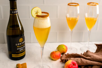 Three champagne flutes filled with a golden drink, garnished with apple slices and cinnamon rims, next to a bottle of sparkling brut and several apples on a marble surface.