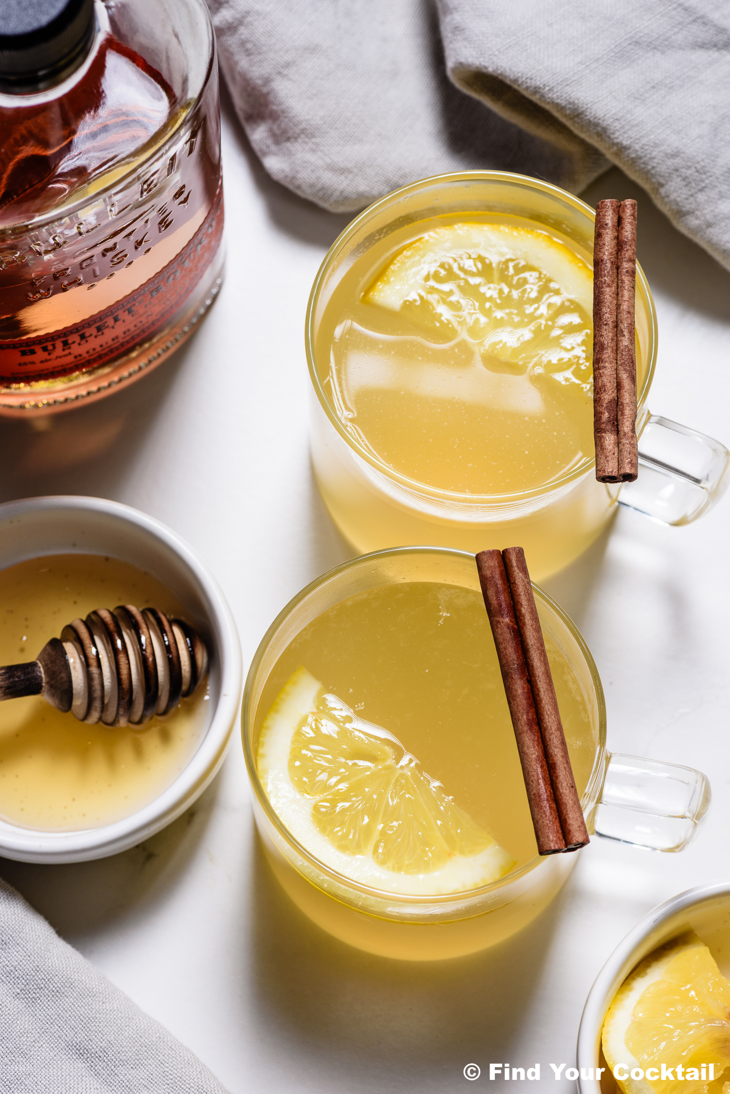 Two glass mugs filled with a yellow drink, each garnished with a lemon slice and cinnamon stick. Nearby are a bottle of whiskey, a bowl of honey with a dipper, and a light cloth.