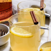 Two clear glass mugs filled with a hot toddy drink, each garnished with a lemon slice and a cinnamon stick, sit next to a bottle of bourbon and a bowl of lemon wedges.