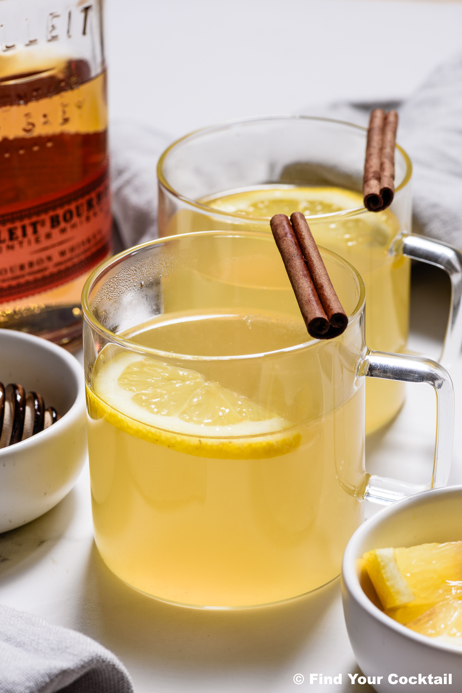 Two clear glass mugs filled with a hot toddy drink, each garnished with a lemon slice and a cinnamon stick, sit next to a bottle of bourbon and a bowl of lemon wedges.