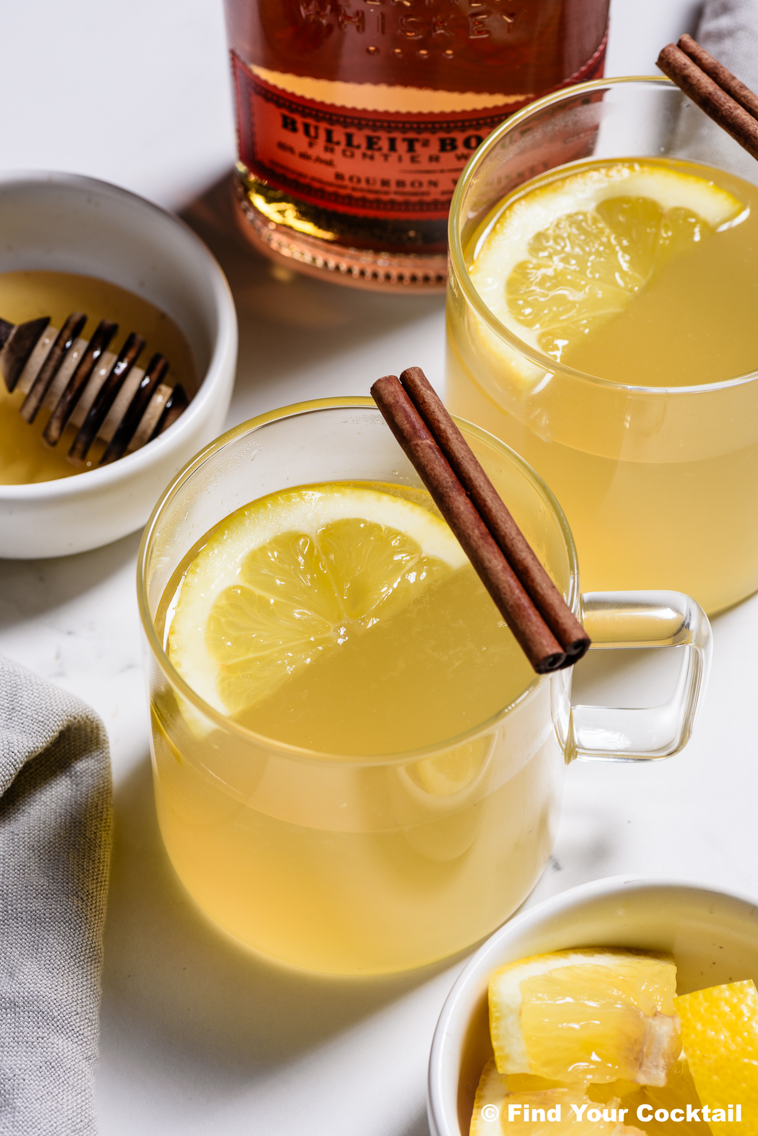 Two glass mugs of hot toddy garnished with lemon slices and cinnamon sticks, with a honey dipper, lemon wedges, and a bottle of bourbon in the background.