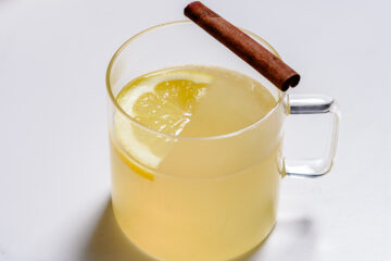 A clear glass mug filled with a light yellow beverage, garnished with a lemon slice and a cinnamon stick on the rim, on a white background.