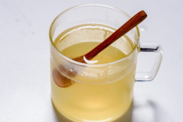 A clear glass mug filled with light yellow liquid, with a wooden spoon resting inside, on a white surface.