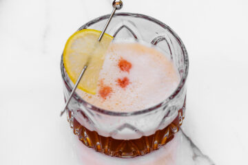 A cocktail in a faceted glass with a lemon slice garnish and foam on top, decorated with three drops of bitters, on a white surface.