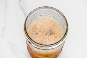 A close-up, top-down view of a glass jar filled with a frothy, light brown liquid on a white surface.