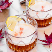 Two whiskey sour cocktails with frothy tops, garnished with lemon slices on cocktail picks, sit on a white surface with red autumn leaves scattered nearby.
