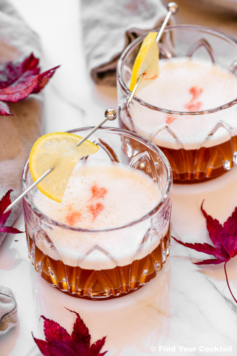 Two whiskey sour cocktails with frothy tops, garnished with lemon slices on cocktail picks, sit on a white surface with red autumn leaves scattered nearby.